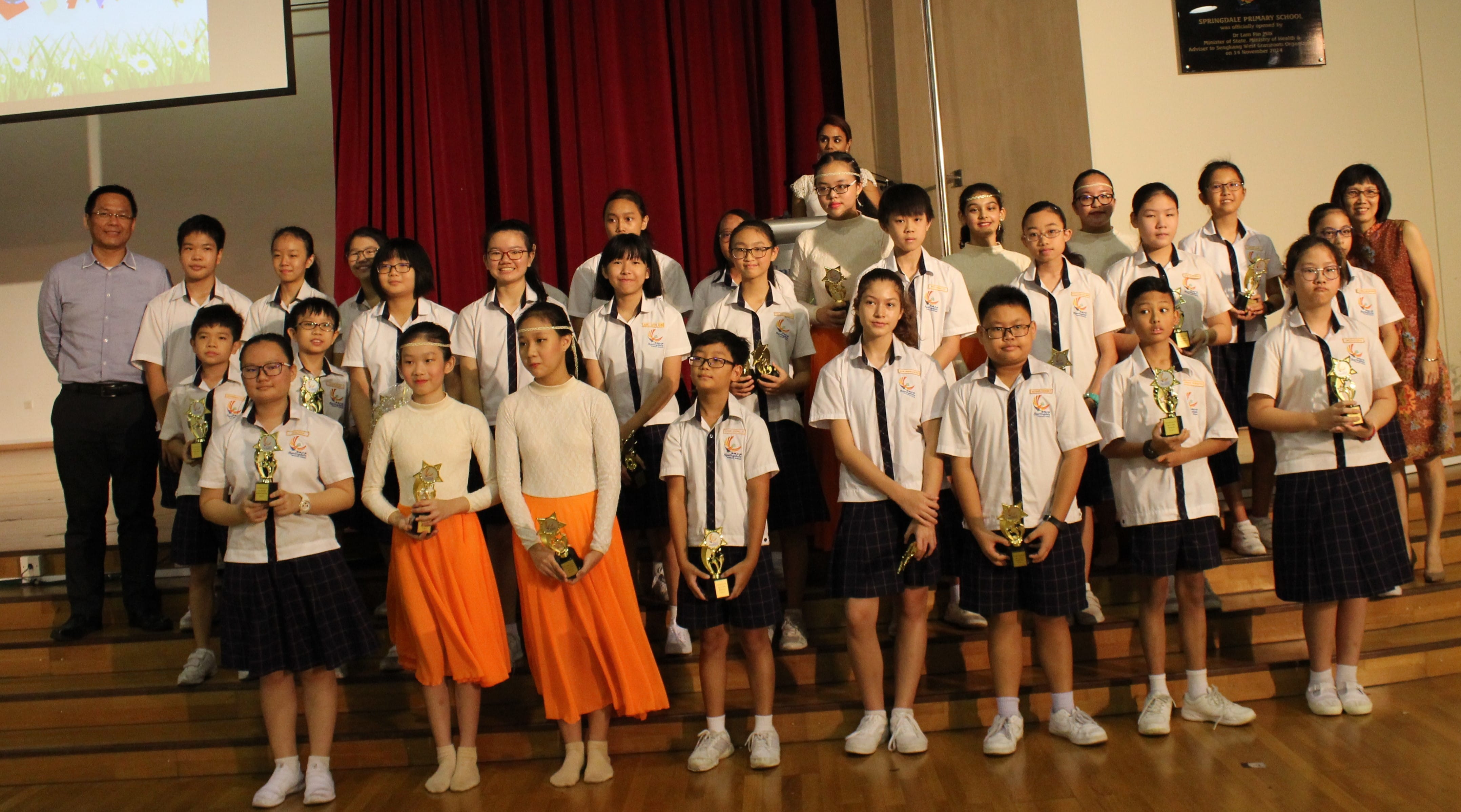 A large group of school students in uniforms holding gold trophies pose on a stage with adults behind them.